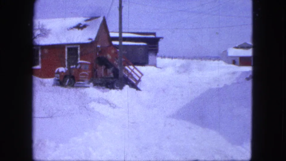 1963 Proctor, Minnesota frame with a red tractor near a red outbuilding, deep snow, utility lines, and distant structures.