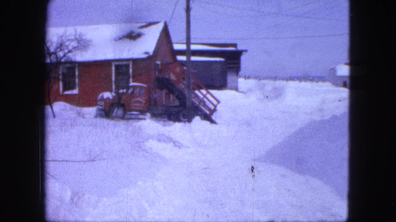 Snow-covered red farm building with stairs, parked vehicle, and utility pole in a rural yard.