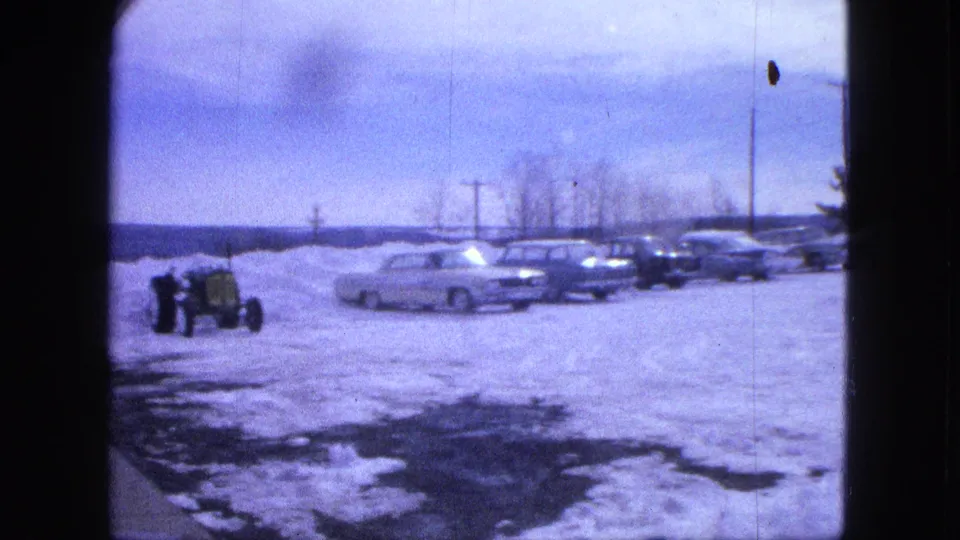 1963 Proctor, Minnesota home-movie frame with a tractor at left and a row of early-1960s cars parked in deep snow.