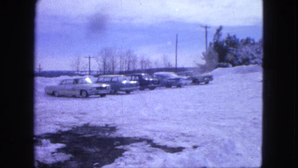1963 Proctor, Minnesota home-movie frame showing several parked cars along a snowy lot with utility poles and trees behind them.