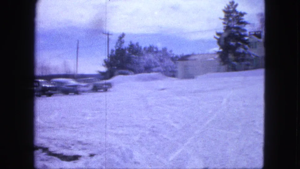 1963 Proctor, Minnesota home-movie frame showing cars leaving the edge of view as the pan reveals snow, a small building, and trees.