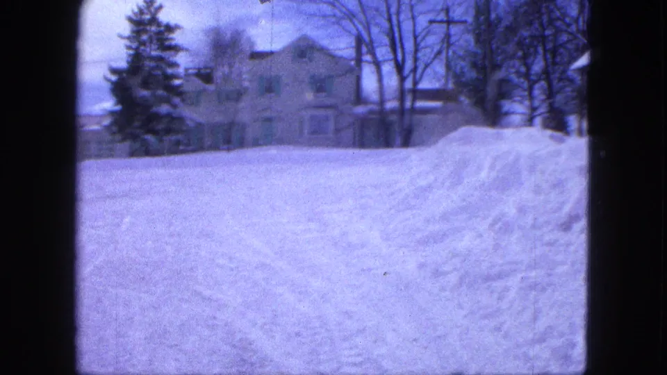 1963 Proctor, Minnesota 8mm frame showing a snowbank beside a residential road with a large house and winter trees beyond.