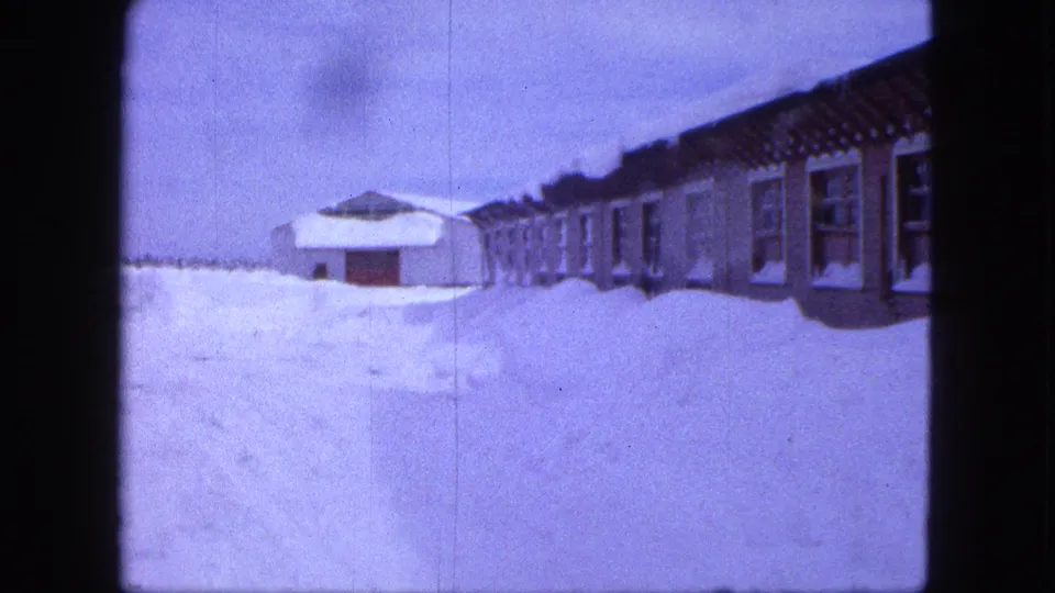 1963 Proctor, Minnesota 8mm frame showing a long building with repeated windows beside deep snow and a smaller structure in the distance.