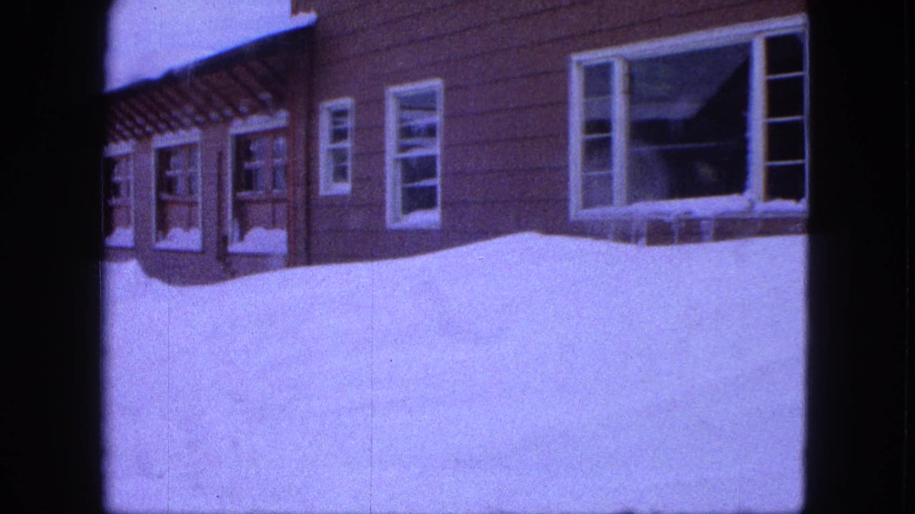 Snowbank rises against a brown-sided house with partially visible windows.