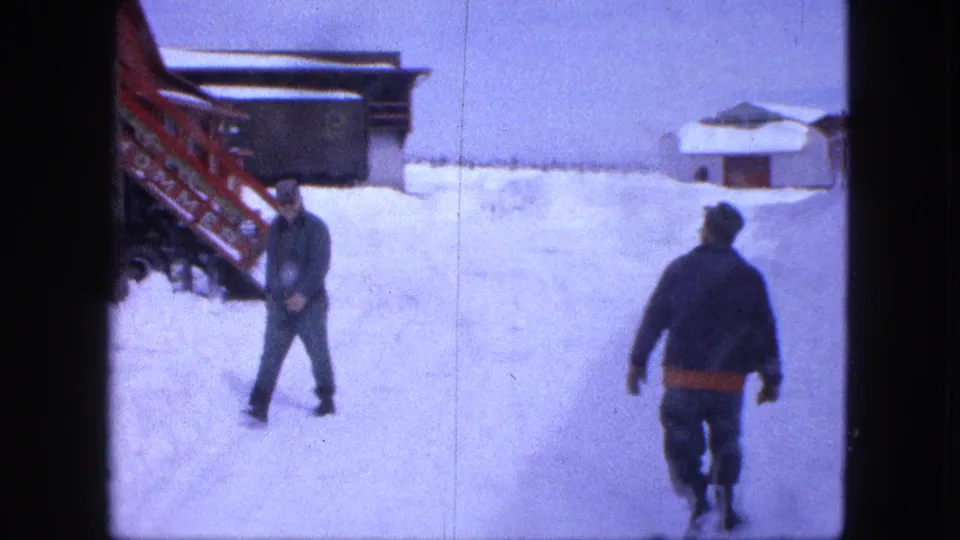 A rear and side view of two men crossing a deep snowy residential yard in 1963 Proctor, Minnesota footage.