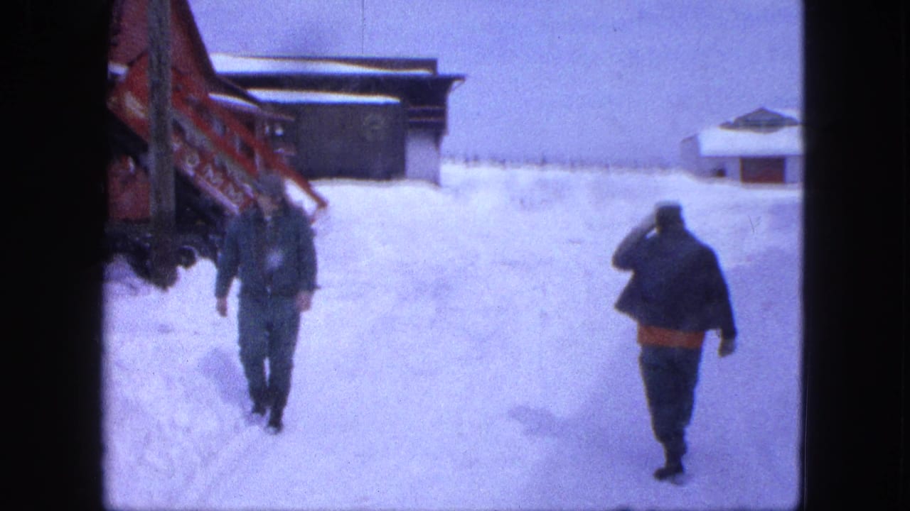 Two men walk through deep snow near a red structure and low buildings.