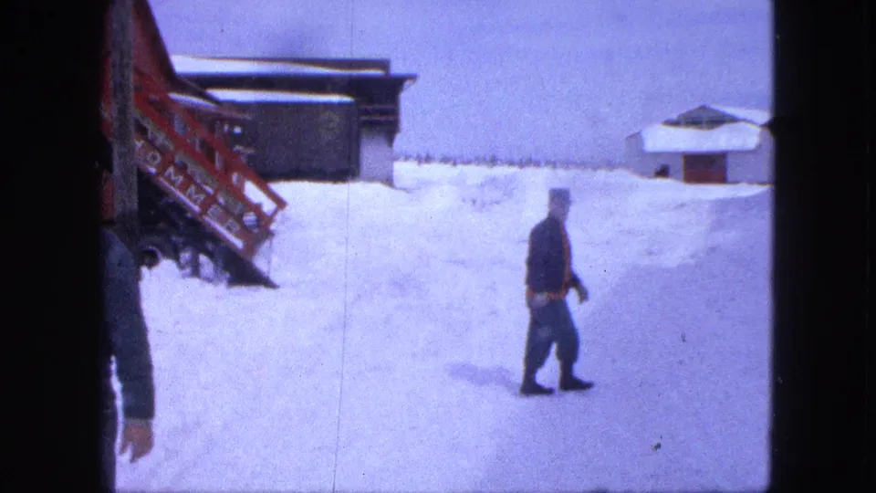One man exits the left edge as another walks through the snow near a red structure and low building in 1963 Proctor home-movie footage.
