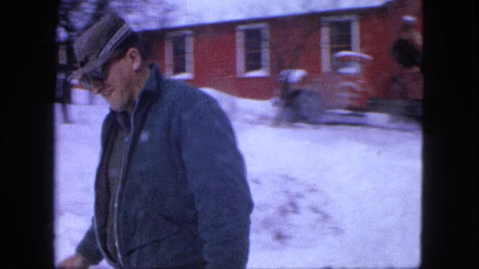 A man wearing a cap and dark jacket passes near the camera, with a red building and snowbanks behind him in Proctor, Minnesota.