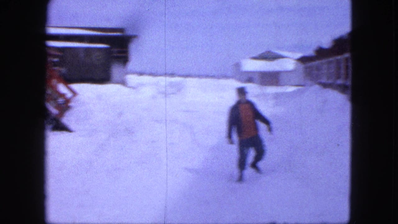 A bundled man moves across deep snow in 1963 Proctor, Minnesota, with low buildings and snow-covered roofs behind him.