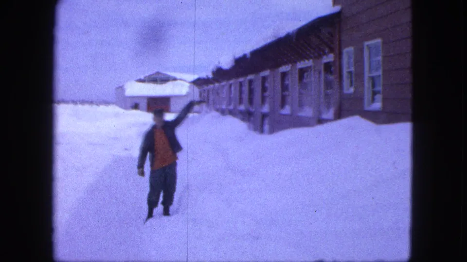 A man in a dark coat and red vest or shirt gestures in deep snow beside a row of snow-covered buildings in Proctor, Minnesota.