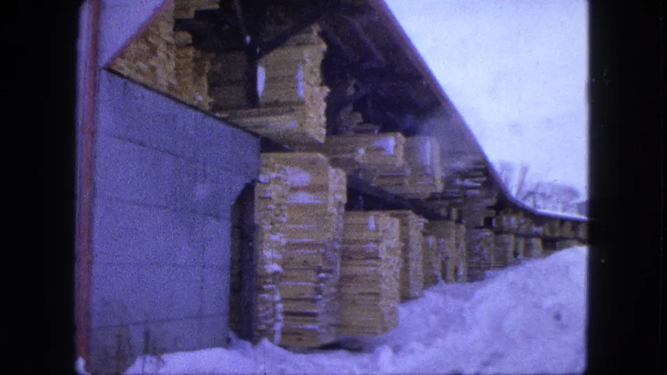 Stacks of lumber are stored under a shelter beside a snow-covered building in this 1963 Proctor, Minnesota home-movie frame.