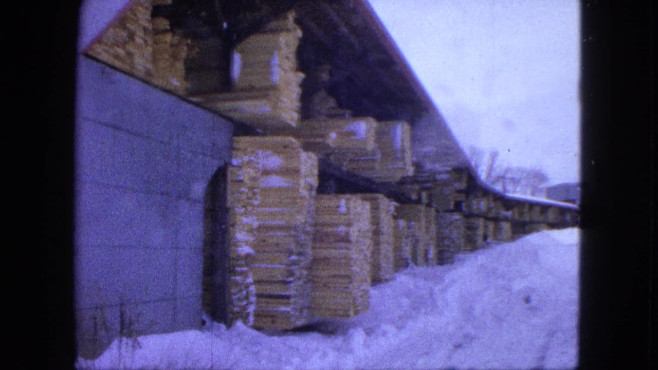1963 Proctor, Minnesota 8mm frame showing stacked lumber under an open shelter with deep snow along the yard path.