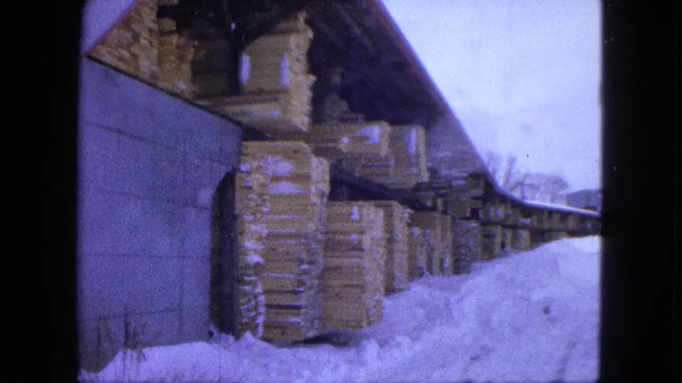1963 Proctor, Minnesota 8mm frame showing stacked lumber under an open shelter with deep snow along the yard path.
