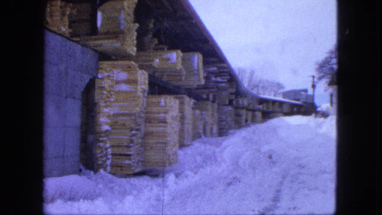 Moving 8mm view of a Proctor lumberyard in winter, with snow piled beside covered stacks of lumber.