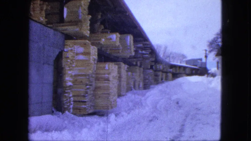Moving 8mm view of a Proctor lumberyard in winter, with snow piled beside covered stacks of lumber.
