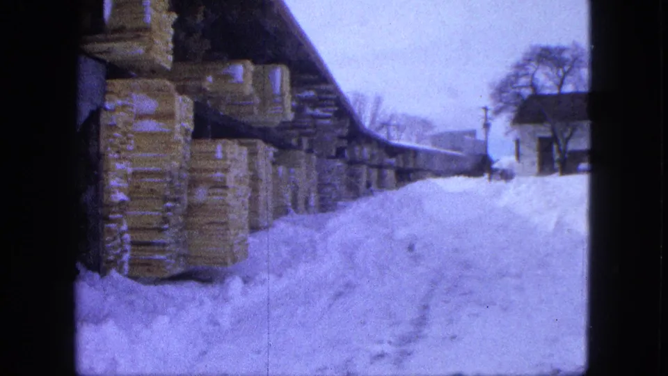 1963 home-movie frame from Proctor showing a snow-covered yard, lumber stacks, a small building, and winter trees.
