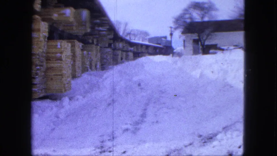 End frame of the Proctor winter lumberyard shot, with stacked boards, deep snow, and a building at right.