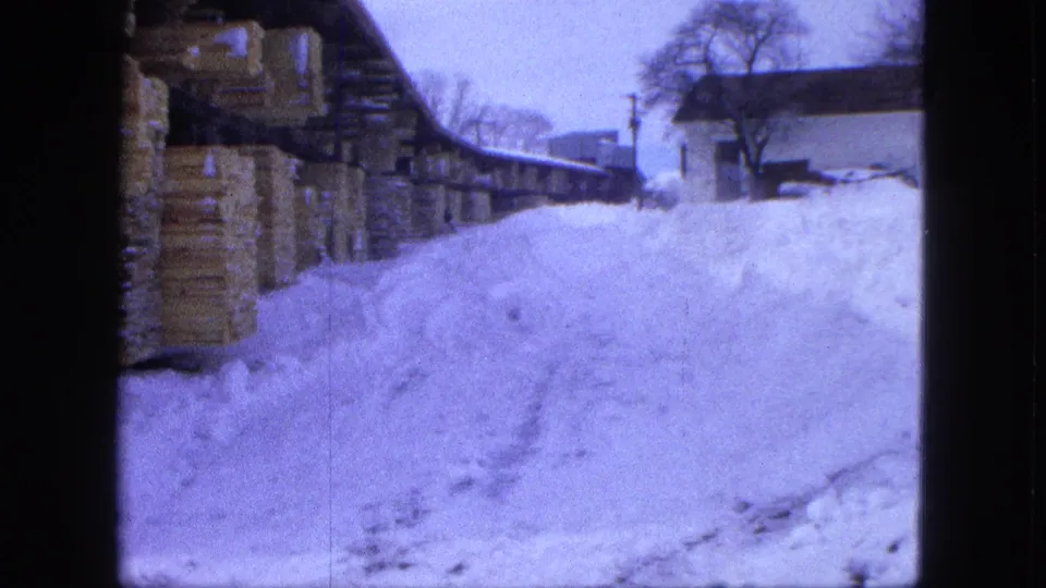 1963 Proctor, Minnesota 8mm frame showing stacked lumber under a shed with heavy snowbanks and bare trees beyond.