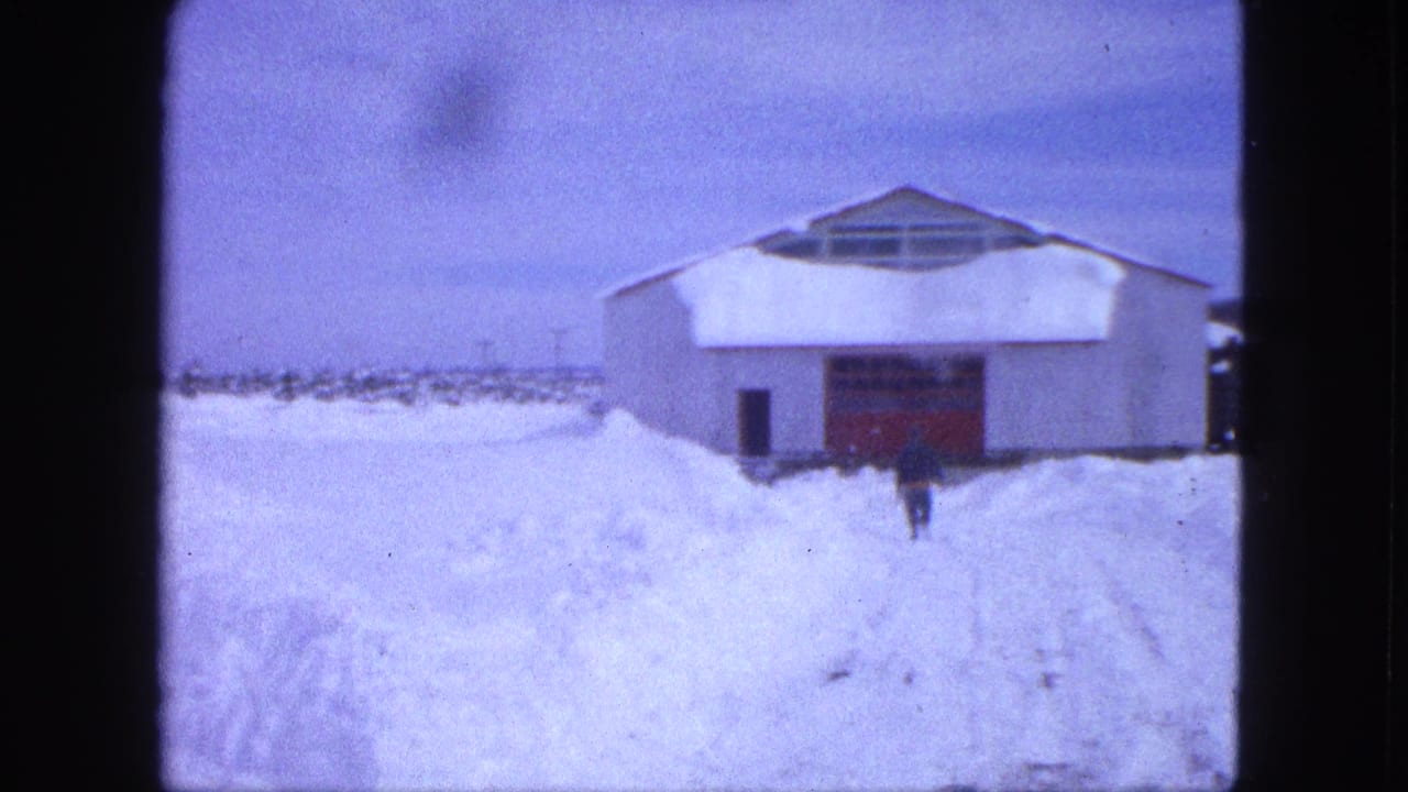 A small figure moves across snow in front of a white building with a red door.