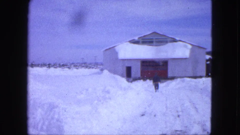 A solitary figure continues toward the red-doored white building in snowy Proctor, Minnesota, seen in soft 8mm color.