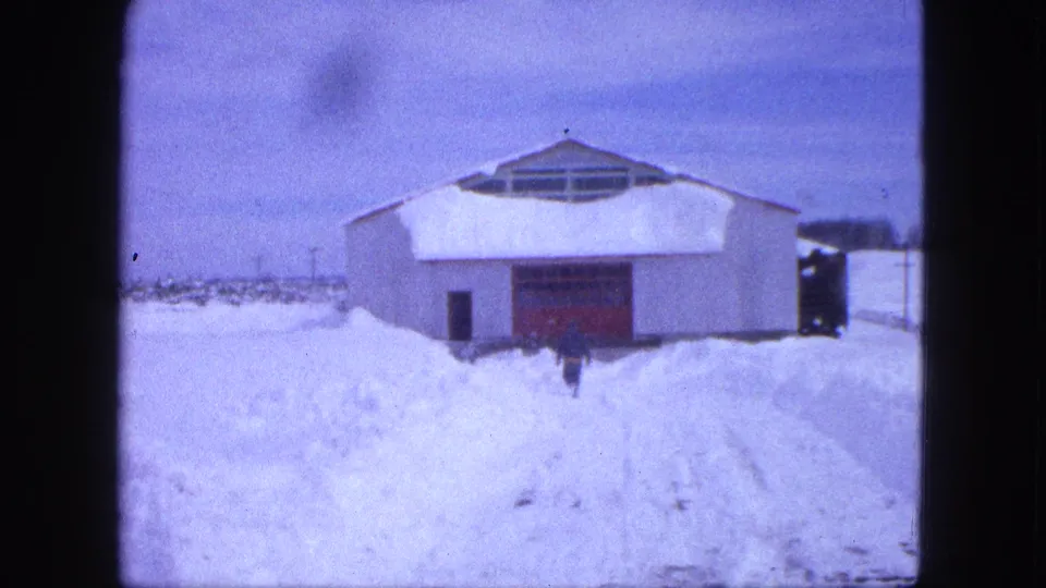 The figure is closer to the white building and red door, with deep snowbanks and plowed tracks visible in the foreground.