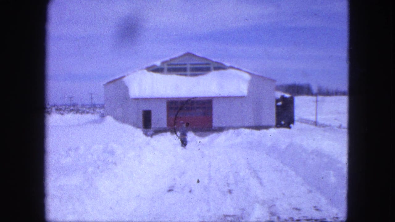 1963 Proctor, Minnesota 8mm frame showing a snowbound white barn-like building and a distant figure near the red door.