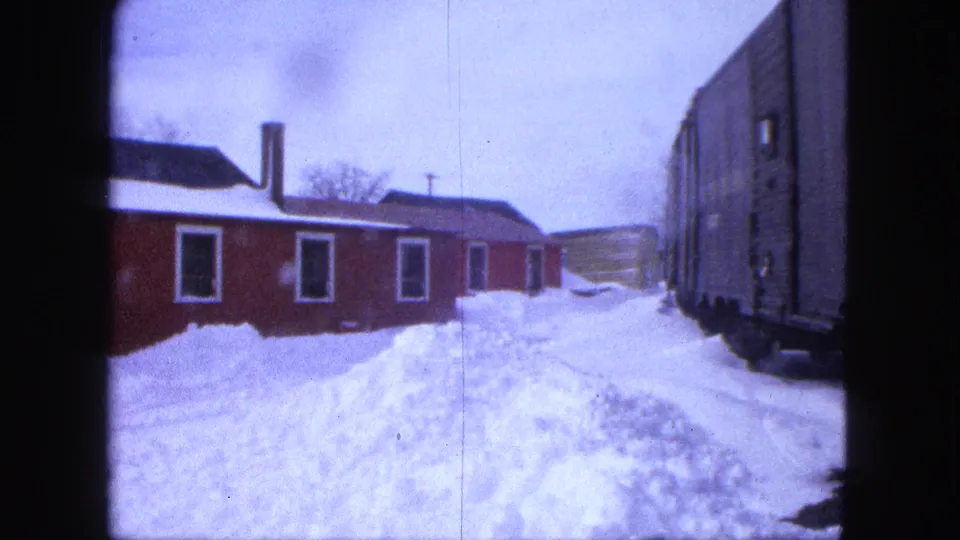 Handheld 8mm view in Proctor, Minnesota showing snow piled between a red building and a rail car in 1963.