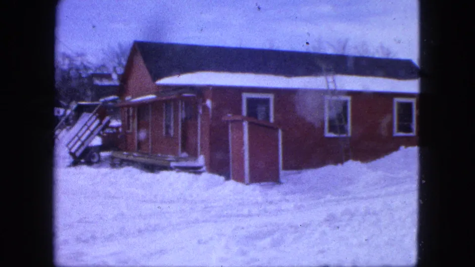 1963 Proctor, Minnesota 8mm frame showing the side of a red house, exterior steps, a small shed, and deep snow in the yard.