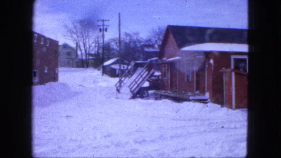 1963 Proctor, Minnesota winter frame showing a snowy yard beside a red house, with exterior steps, a shed, bare trees, and utility poles.
