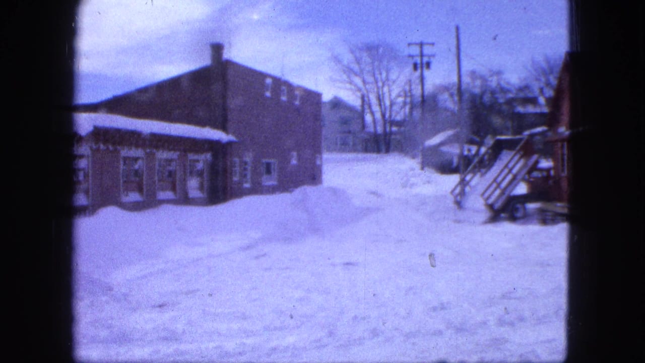 1963 Proctor, Minnesota home-movie frame with lingering snow, brick buildings, a porch edge, bare trees, and utility poles.