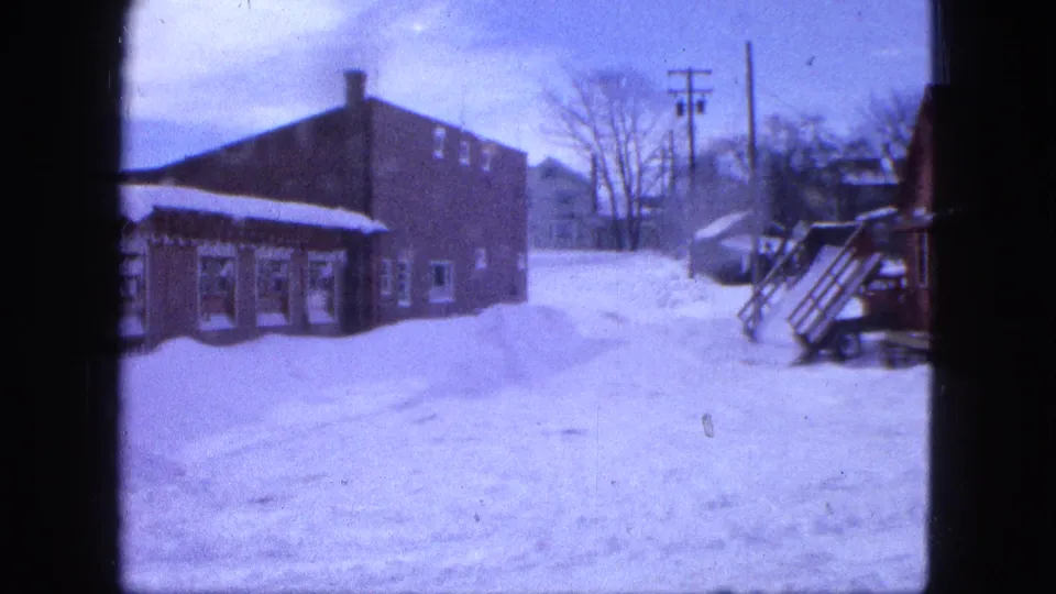 1963 Proctor, Minnesota home-movie frame with lingering snow, brick buildings, a porch edge, bare trees, and utility poles.