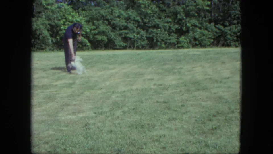 A man in dark clothing bends down toward a small white dog on grass in Saint Paul, Minnesota, in 1963 home-movie footage.