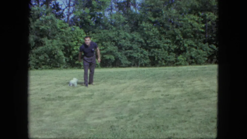 A man in dark clothing stands on the grass with the small white dog close by in the 1963 Saint Paul backyard clip.