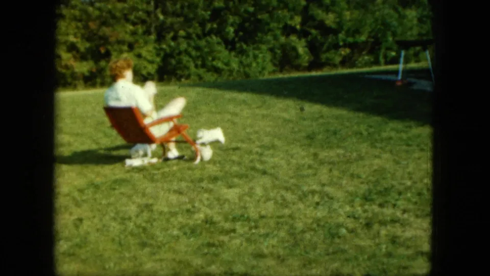 Home-movie frame from Saint Paul in 1963 showing a seated woman and small white dogs in a suburban backyard.