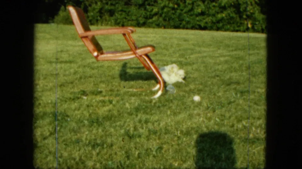 1963 Saint Paul home-movie still showing a small white dog close to a wooden lawn chair, with a ball in the grass nearby.