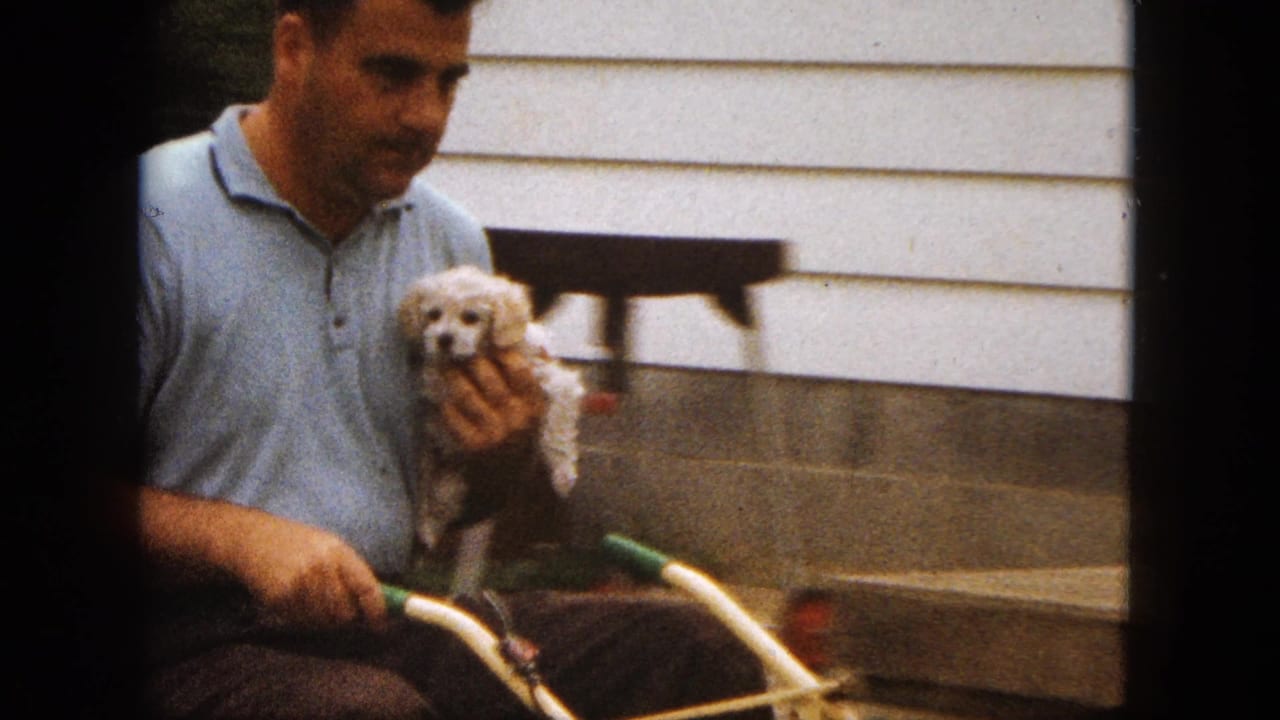 In 1963 Saint Paul home-movie footage, a man in a light blue polo holds a small white poodle puppy near a house exterior and lawnmower handlebars.