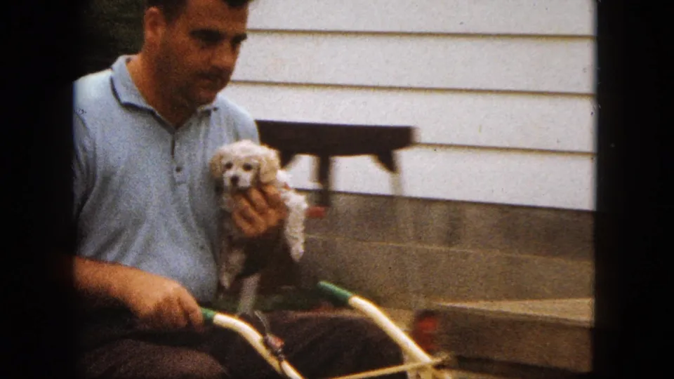 In 1963 Saint Paul home-movie footage, a man in a light blue polo holds a small white poodle puppy near a house exterior and lawnmower handlebars.