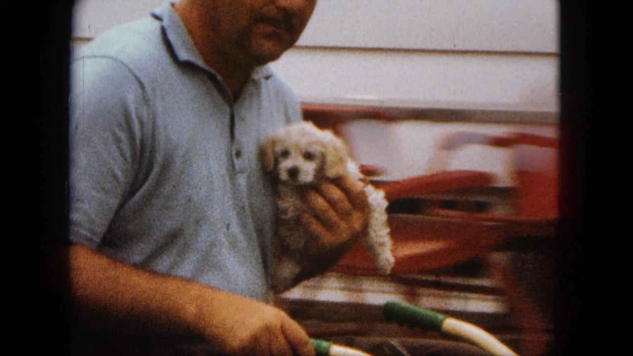 The man holds the small white poodle puppy close to his chest as a red car and house siding appear in the background.