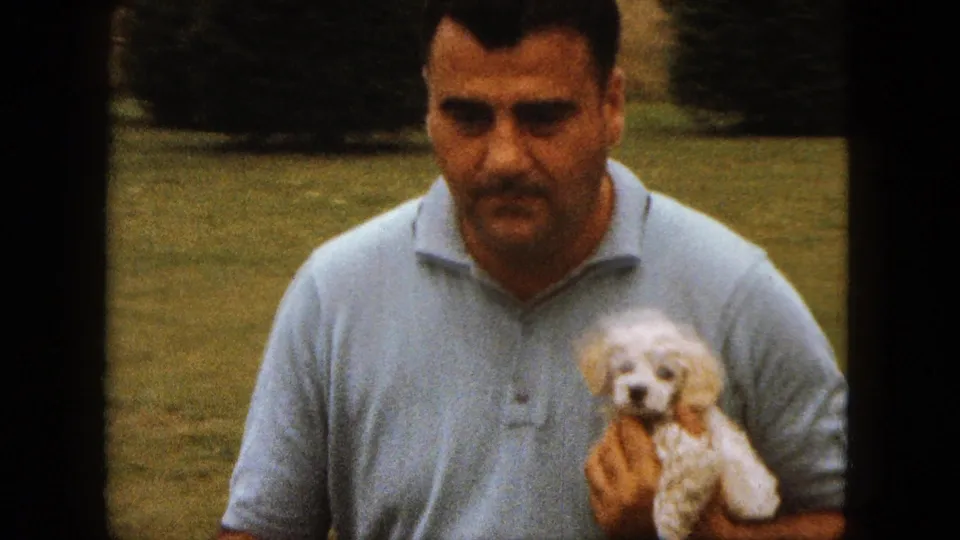 The man faces the camera in a grassy yard while holding the small white poodle puppy, with trees or shrubs behind him.