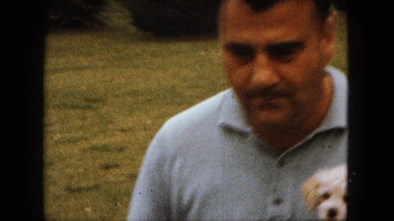 Man in a light blue polo holds a small white dog in a grassy yard.