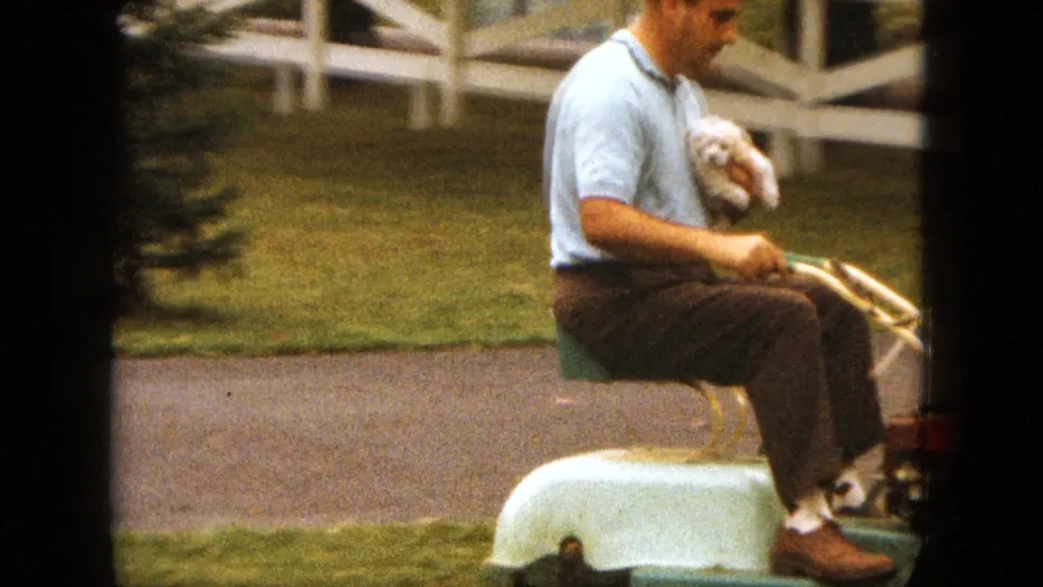 A man in a light blue polo rides a vintage mower in Saint Paul while holding a small white dog near grass and a white fence.