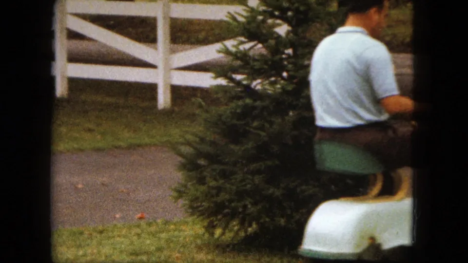 The man continues riding the lawn mower through the 1963 Saint Paul yard, with an evergreen tree, grass, and white fence behind him.