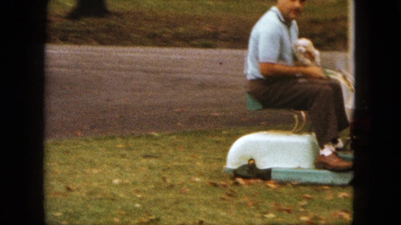 Man sits on a small mower holding a white puppy near grass and a road.