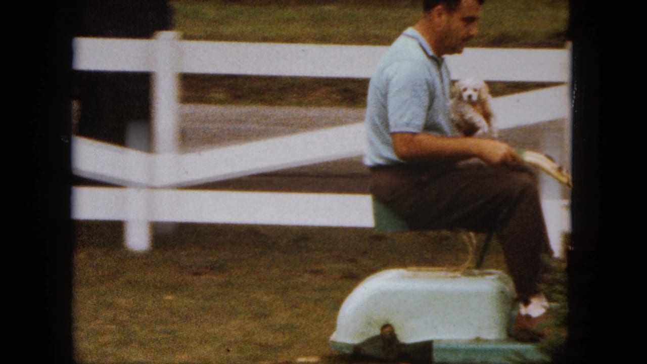Man sitting on a pale ride-on mower while holding a small white puppy near a white fence.