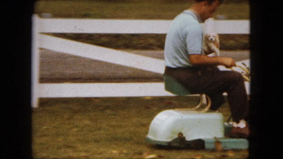 A side view shows the man in a light blue polo seated on the mower, holding the small puppy against a white fence in the Saint Paul yard.