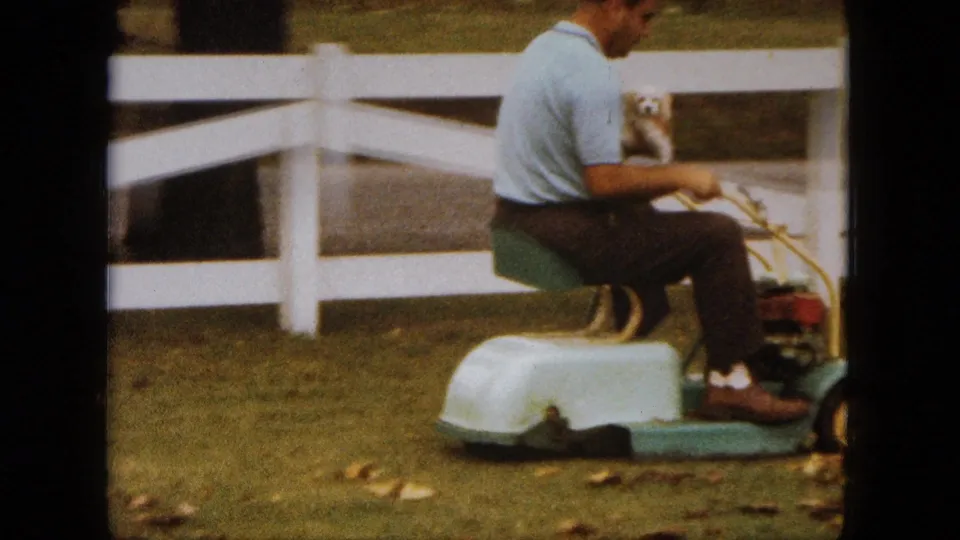 The man remains seated on the vintage ride-on mower with the puppy in his lap, framed by grass, leaves, and a white fence in Saint Paul.