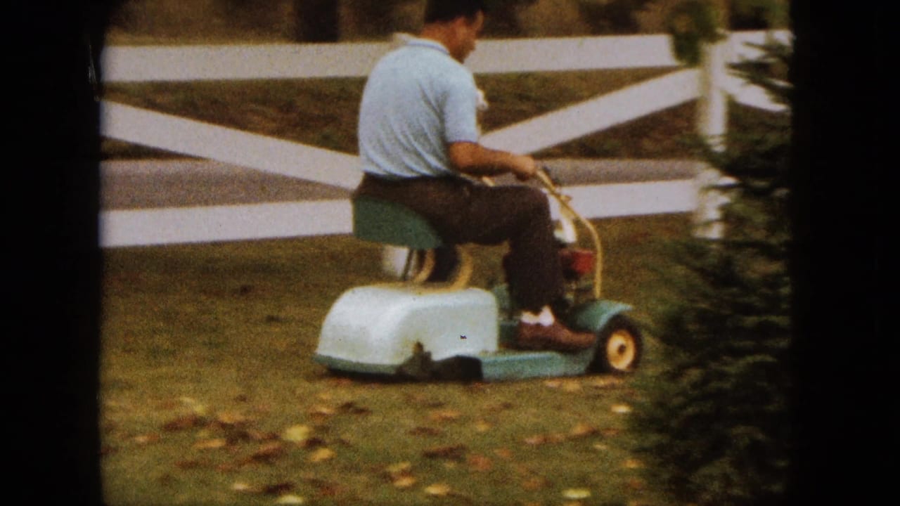 A man in a light blue shirt rides a small mower through a Saint Paul yard, with a white fence and fallen leaves behind him.