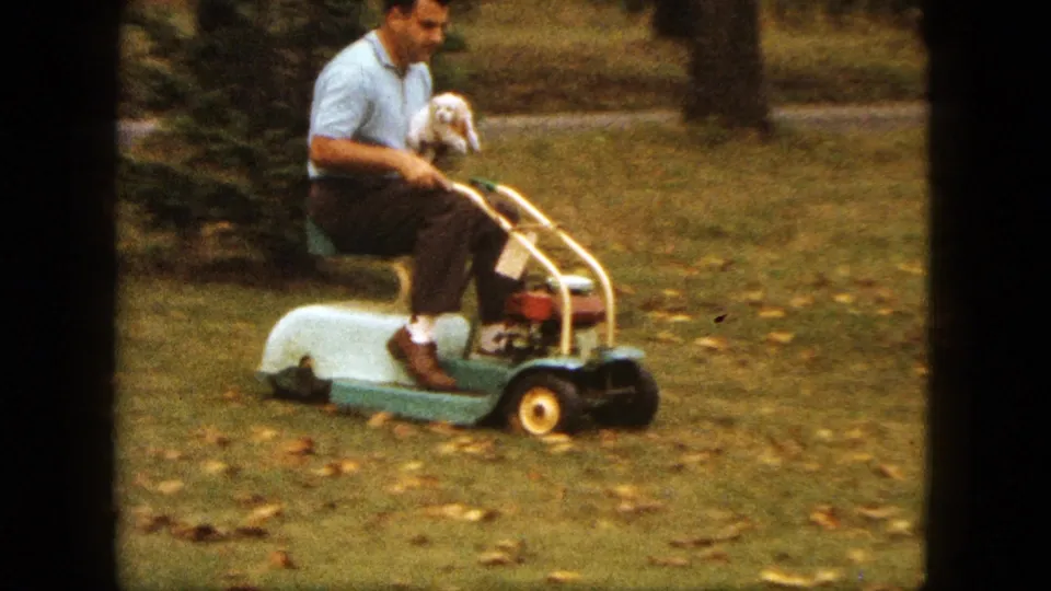 A man smiles while riding a compact green and white lawnmower in a Saint Paul yard, holding a small white dog against his chest.