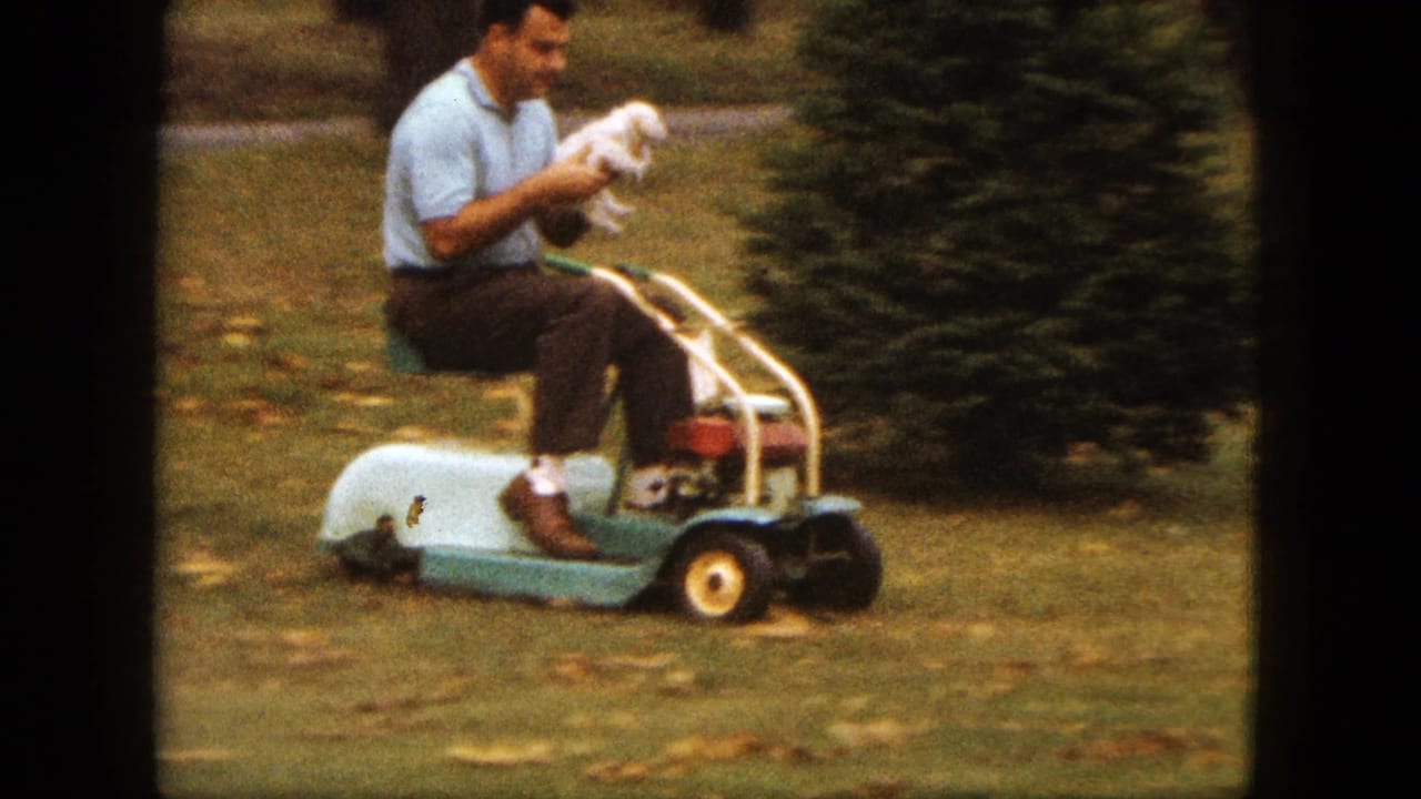 Man holding a small white puppy while seated on a riding lawnmower in a grassy yard.