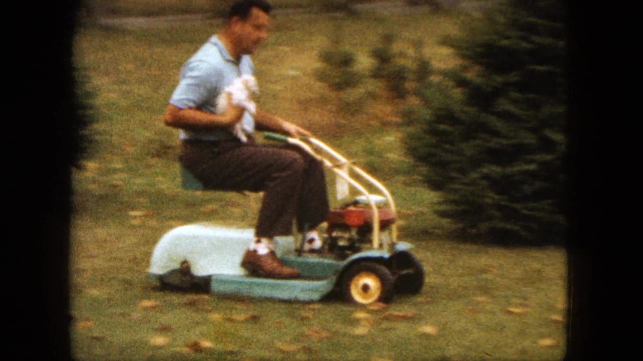 A man in a light blue shirt rides a vintage lawnmower across a Saint Paul yard while cradling a small white puppy.